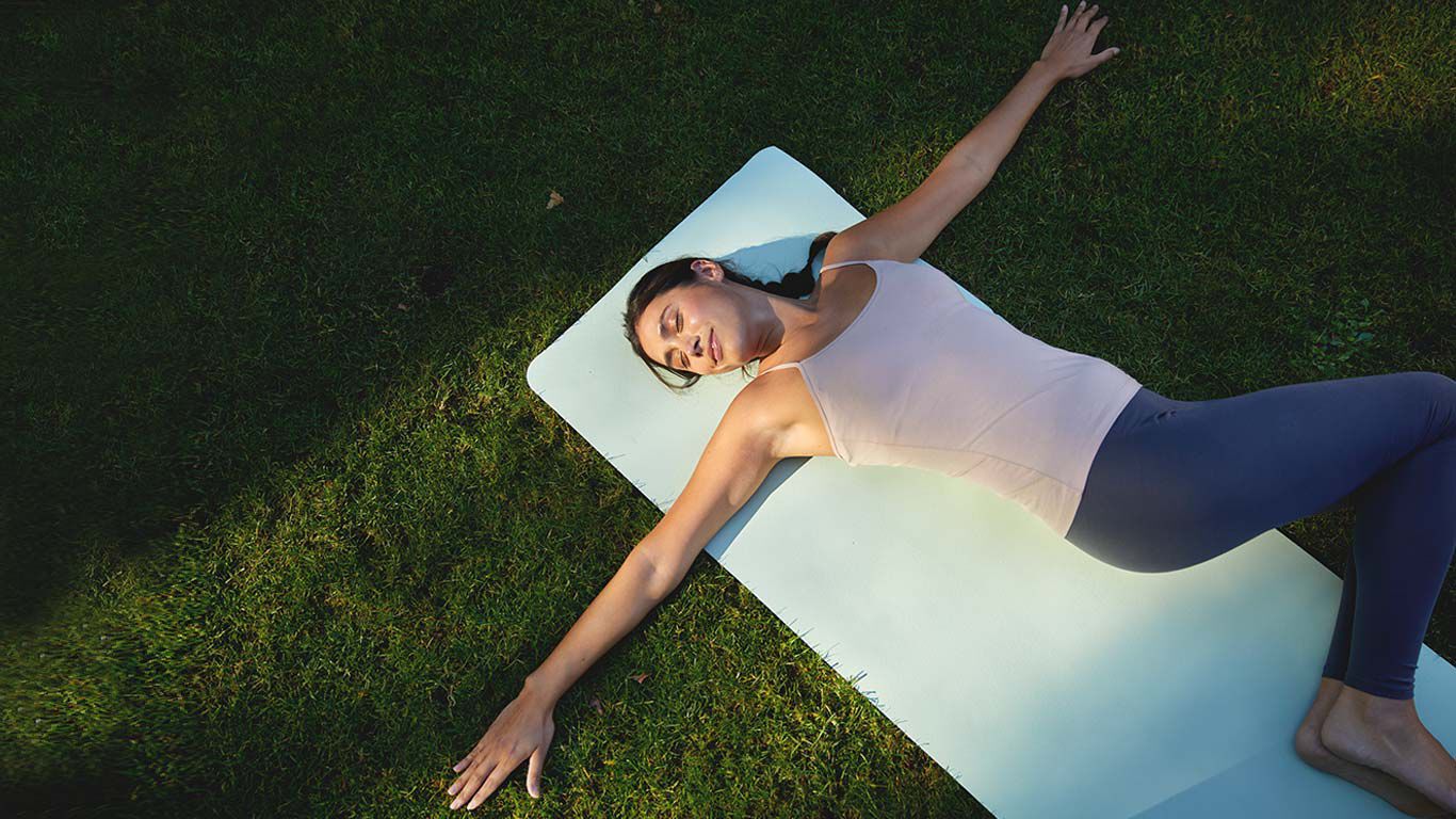 Frau in Trainingsklamotten führt eine liegende Dehnposition auf einer Yogamatte im Gras aus..